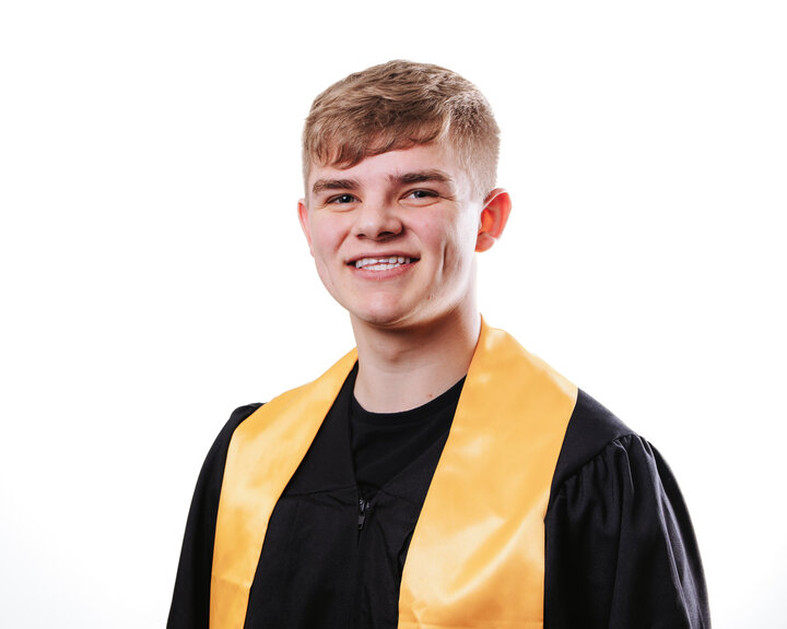 Smiling graduate in black gown with a yellow sash on a white background, Caleb Burnside